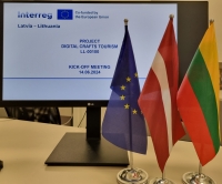 Uz galda stāv ES, Latvijas un Lietuvas karogs, un monitors, uz kura parādīta projekta informācijas kartīte