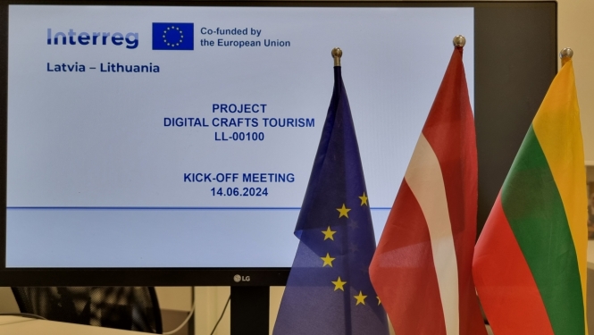 Uz galda stāv ES, Latvijas un Lietuvas karogs, un monitors, uz kura parādīta projekta informācijas kartīte